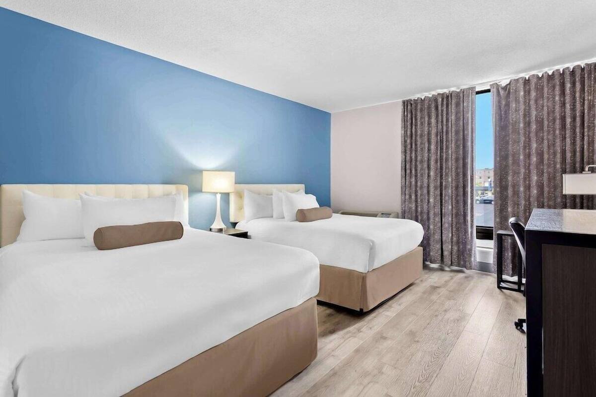 A hotel room with two beds, a desk, a lamp, and a view of the city.