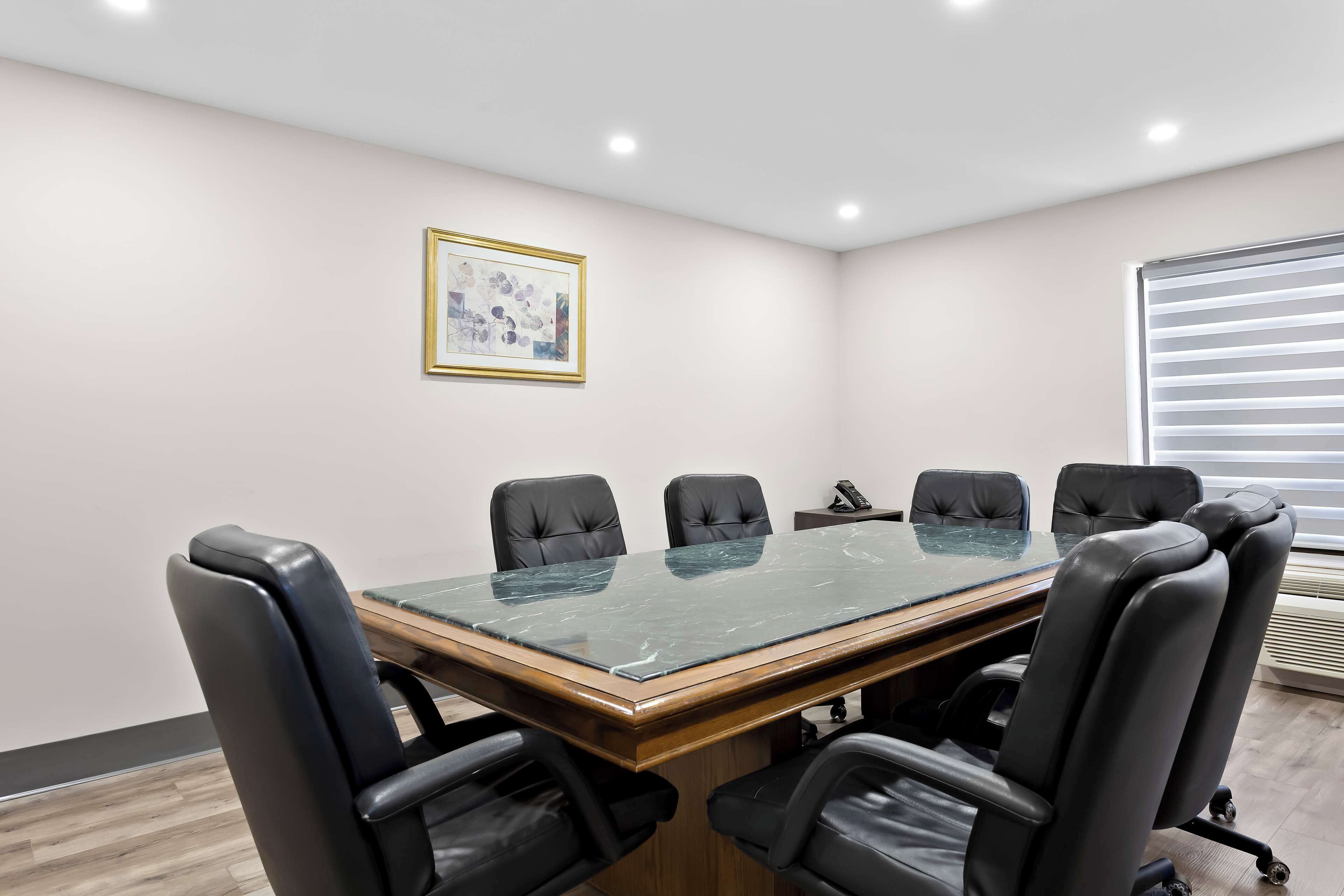 A conference room with a glass-top table, black leather chairs, a framed picture on the wall, and a window with blinds.