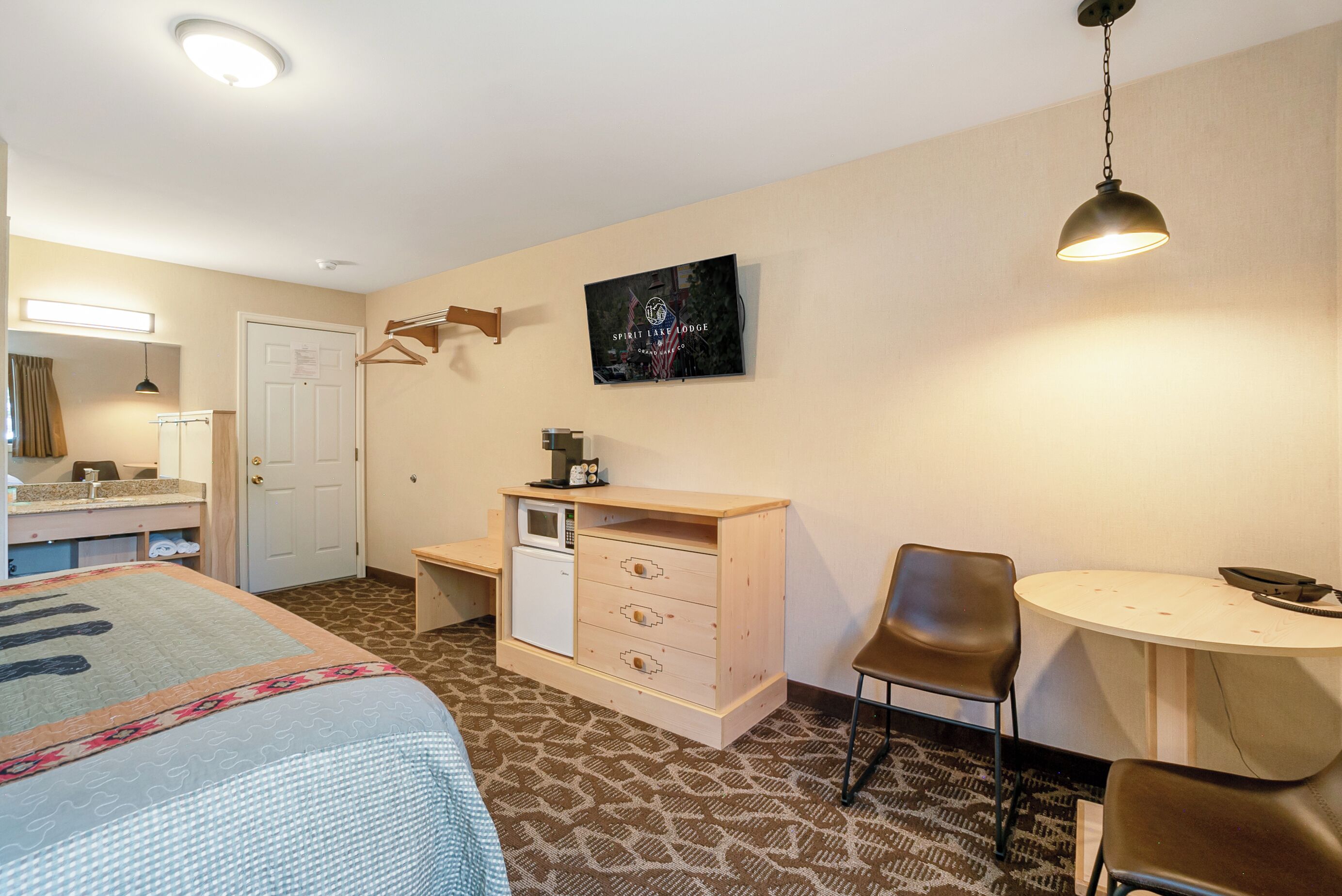 A hotel room with a bed, a desk with a chair, a small table, a microwave, and a TV on the wall.