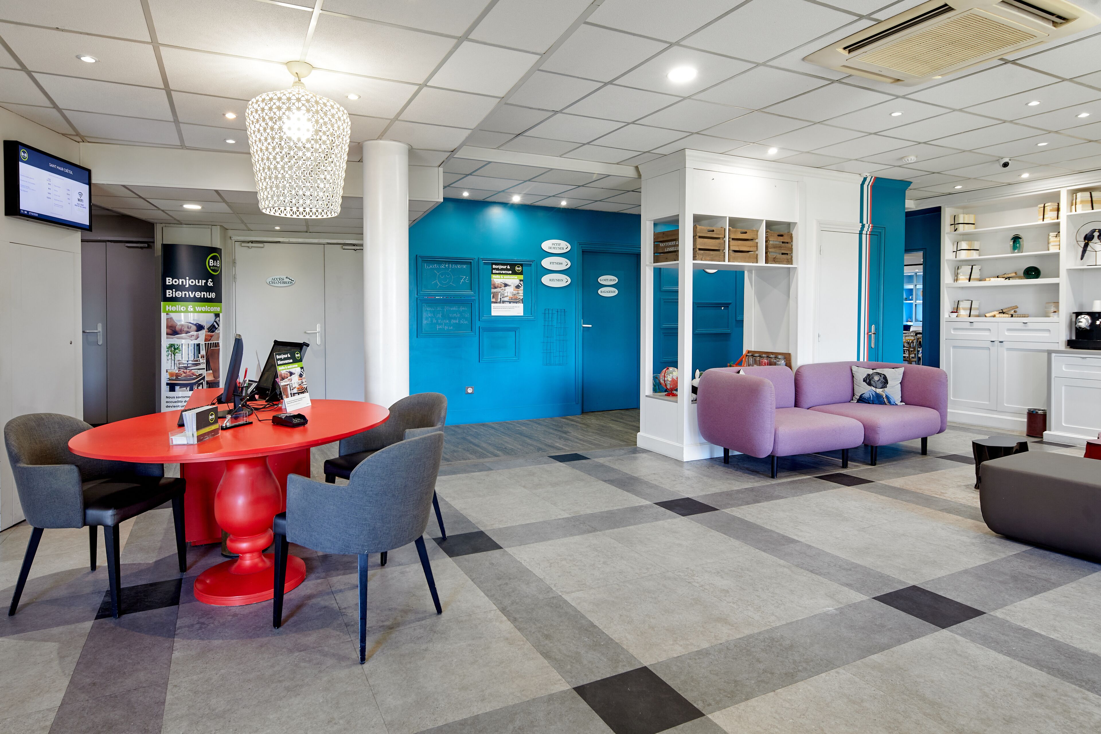 Un lobby moderno con una mesa roja, sillas grises, un sofá morado y una pared azul decorativa.