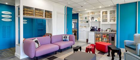 Un salon moderne avec un canapé violet, un pouf gris et un coin cuisine comprenant un réfrigérateur et des placards.