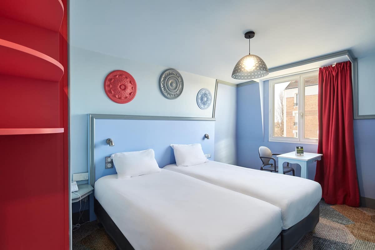 A hotel room with two beds, a desk, a chair, and a window with red curtains.