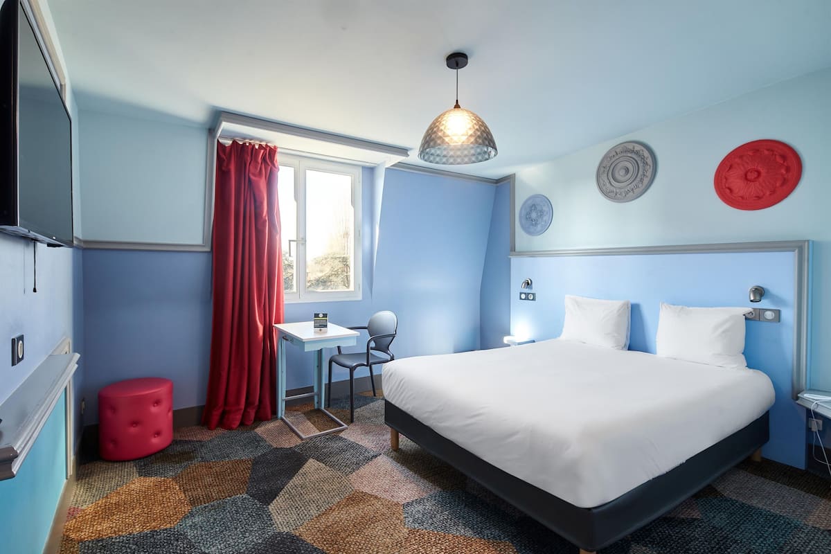 A bedroom with a bed, a desk, a chair, a red stool, a window with red curtains, and decorative plates on the wall.