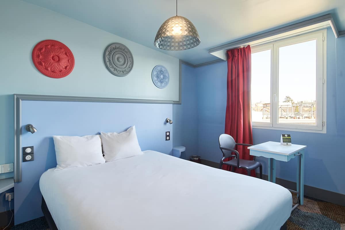 A hotel room with a bed, a desk, a chair, a window with a red curtain, and decorative plates on the wall.