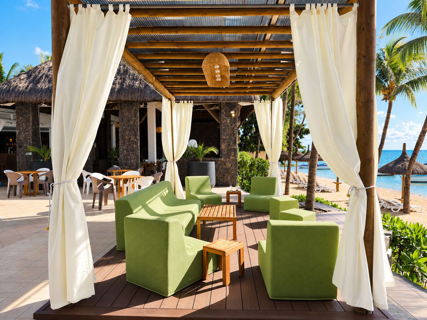 An outdoor lounge area with green cushioned seating, wooden tables, and a thatched-roof structure in the background.