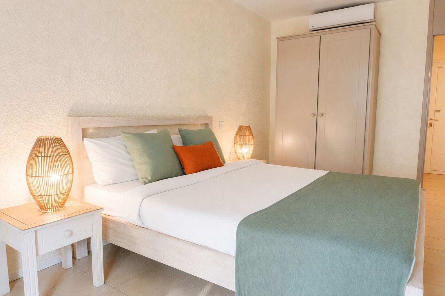 Una cama con cabecera blanca, varios almohadones, una mesita de noche con lámpara y un armario en una habitación con paredes y suelo de tonos claros.
