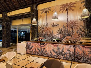 A reception area with a decorative wall featuring palm trees and a fisherman, a counter with a woven lamp, and a tiled floor.