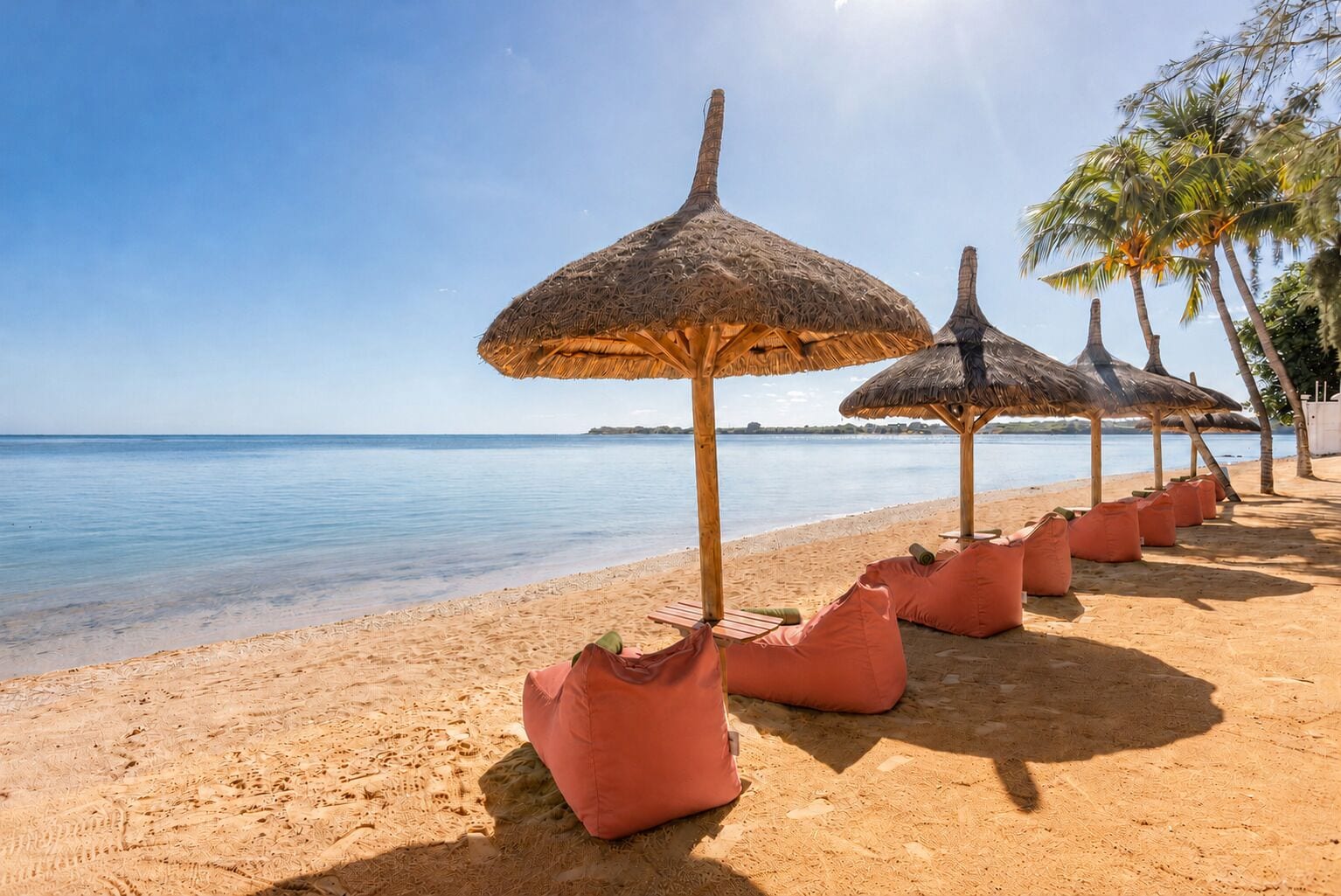 A beach with thatched umbrellas and bean bags, a clear sky, and calm water.