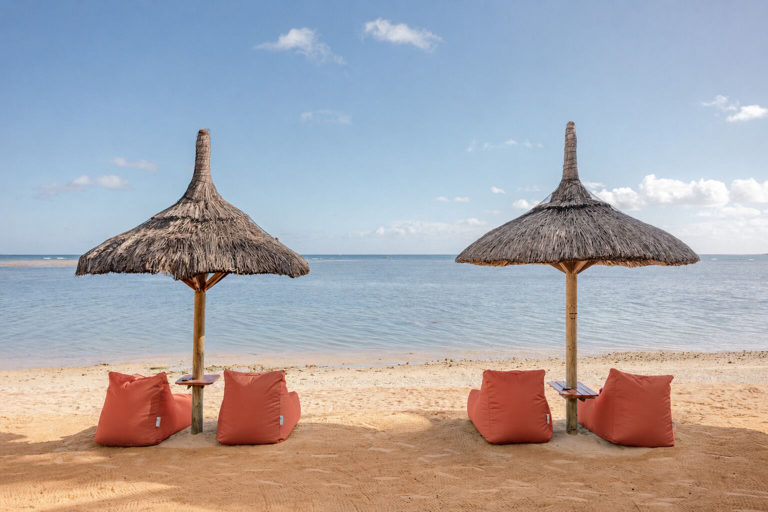 Deux parasols de plage à toits de chaume, agrémentés de coussins, sont installés sur une plage de sable, offrant une vue sur des eaux calmes.