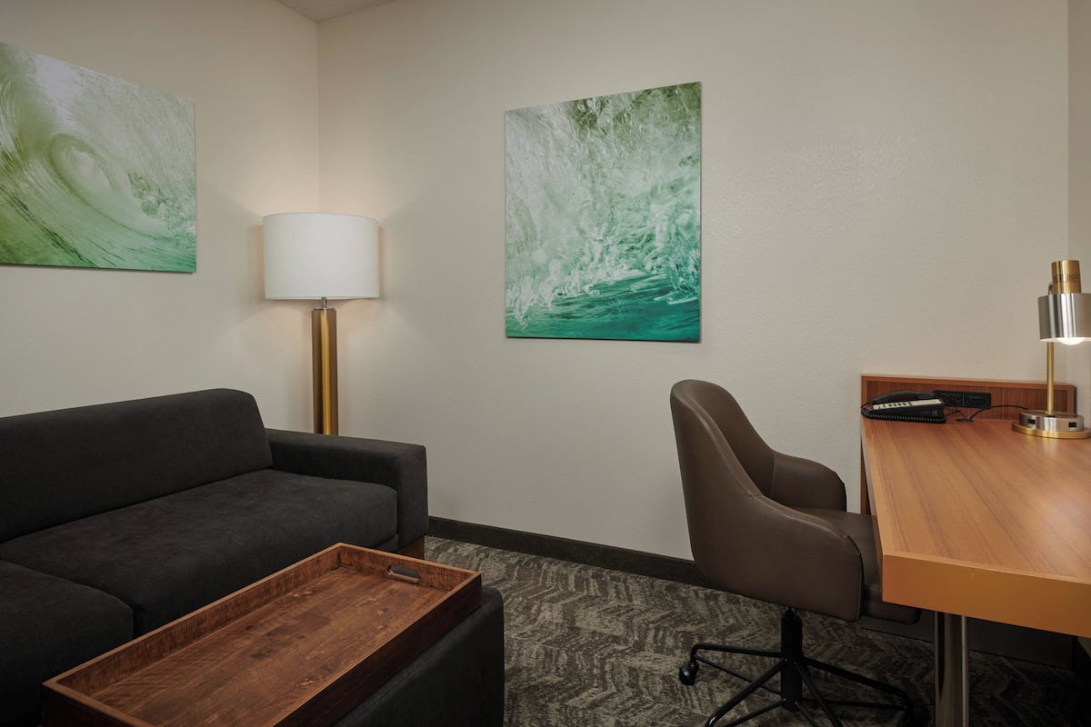 A hotel room with a dark sofa, a wooden coffee table, a desk with a lamp, and two wall-mounted paintings.