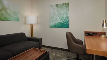 Habitación de hotel con un sofá oscuro, una mesa de centro de madera, un escritorio con lámpara y dos cuadros colgados en la pared.