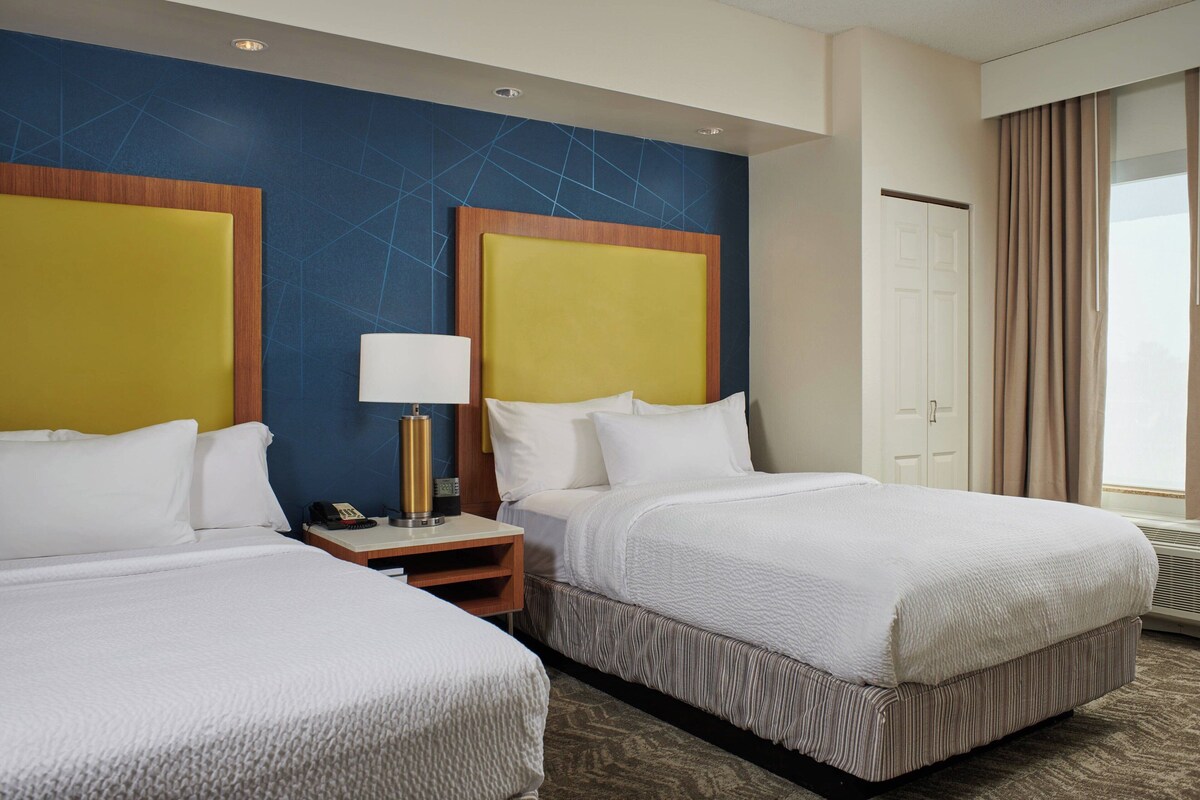 Quarto de hotel com duas camas, mesa de cabeceira, luminária e uma janela grande com cortinas.