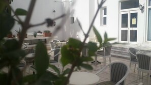 An outdoor seating area with white chairs and tables, potted plants, and a white building with large windows and a door. - Ariha Hotel (Tunis)