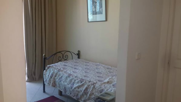 Ein kleines Zimmer mit einem Einzelbett, einer Bettdecke mit Blumenmuster, einem Metallbettgestell, einem roten Teppich und einem gerahmten Bild an der Wand.