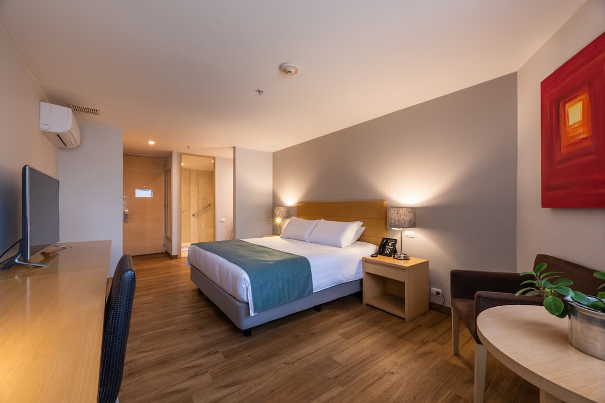 A modern hotel room with a bed, bedside table with a lamp, a desk with a computer, and a chair.