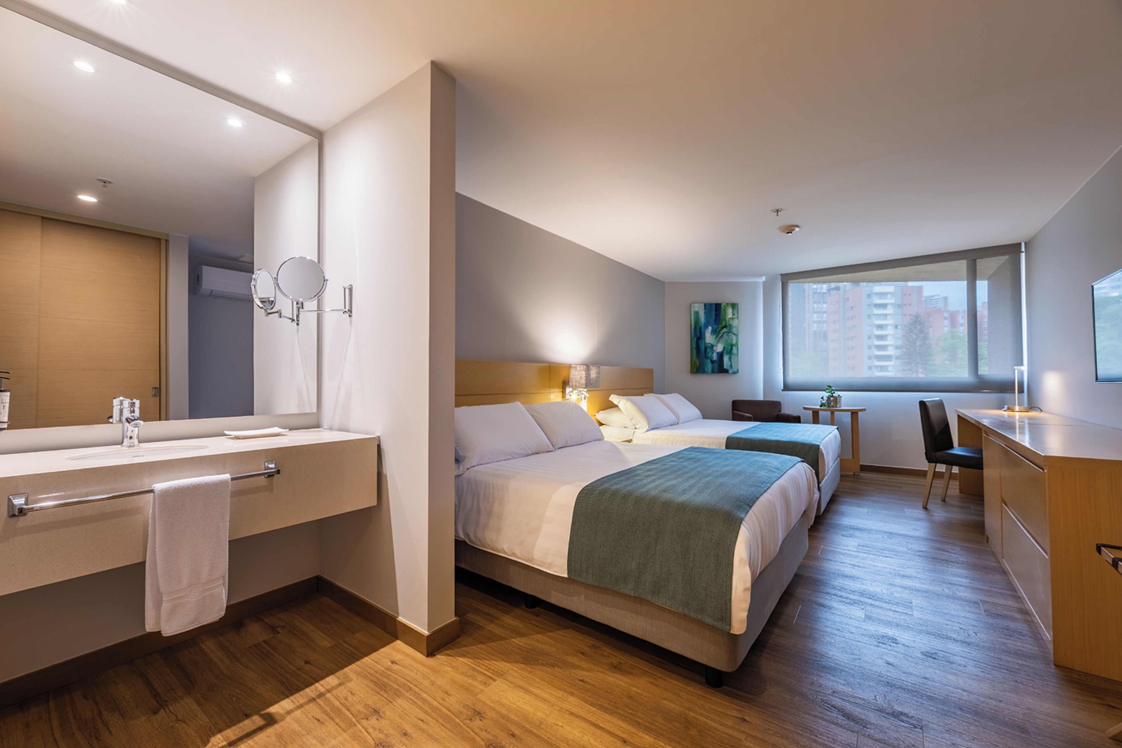 Quarto de hotel com duas camas, uma escrivaninha, uma cadeira, uma mesinha, um espelho e uma janela.