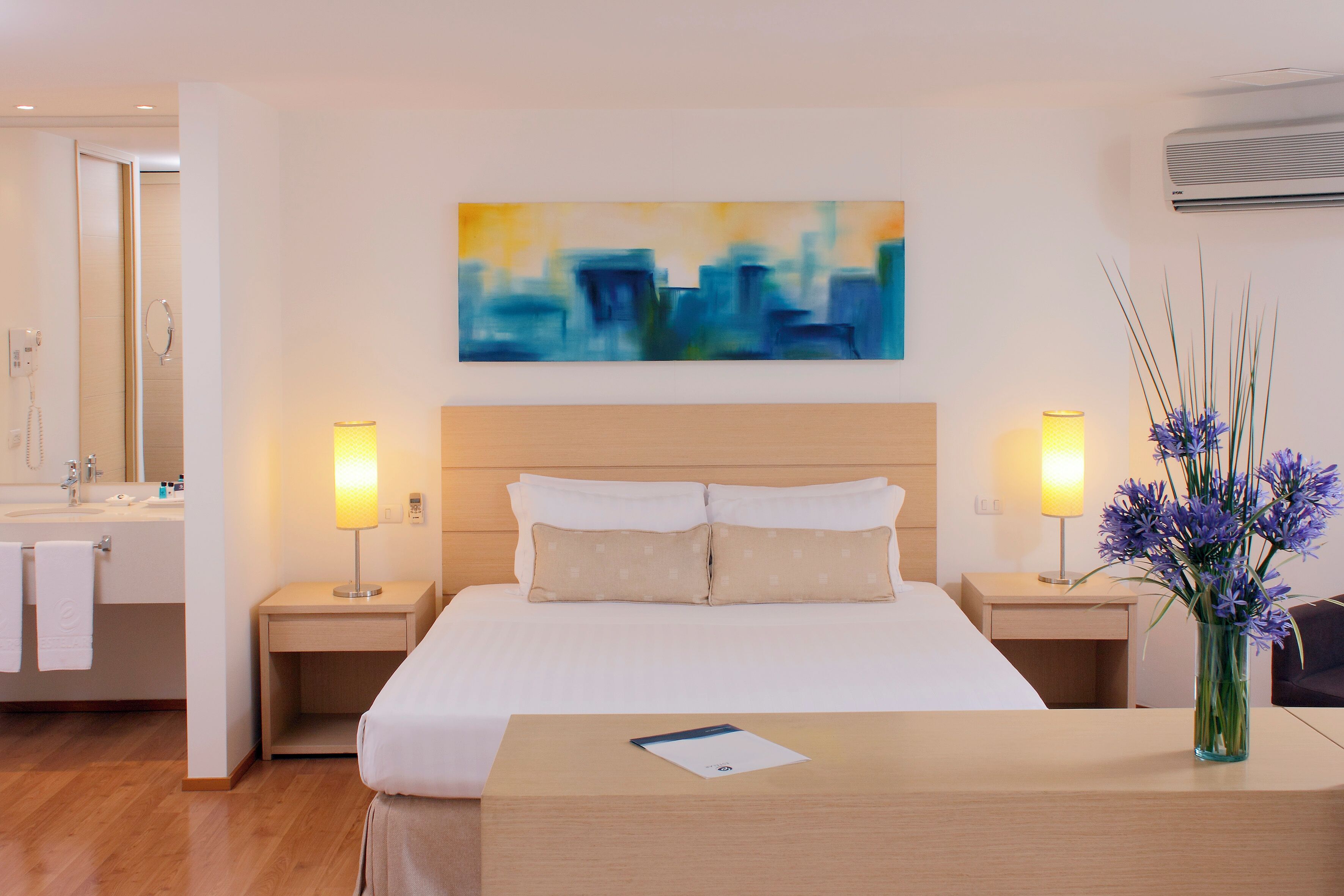Quarto de hotel com cama, mesas de cabeceira, banheiro com pia e espelho, e um quadro na parede.