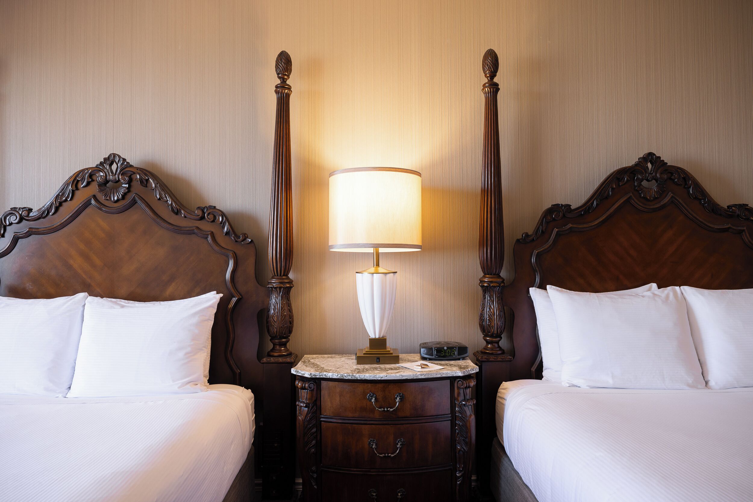 Quarto de hotel com duas camas de madeira, uma mesa de cabeceira com abajur e uma gavetinha.