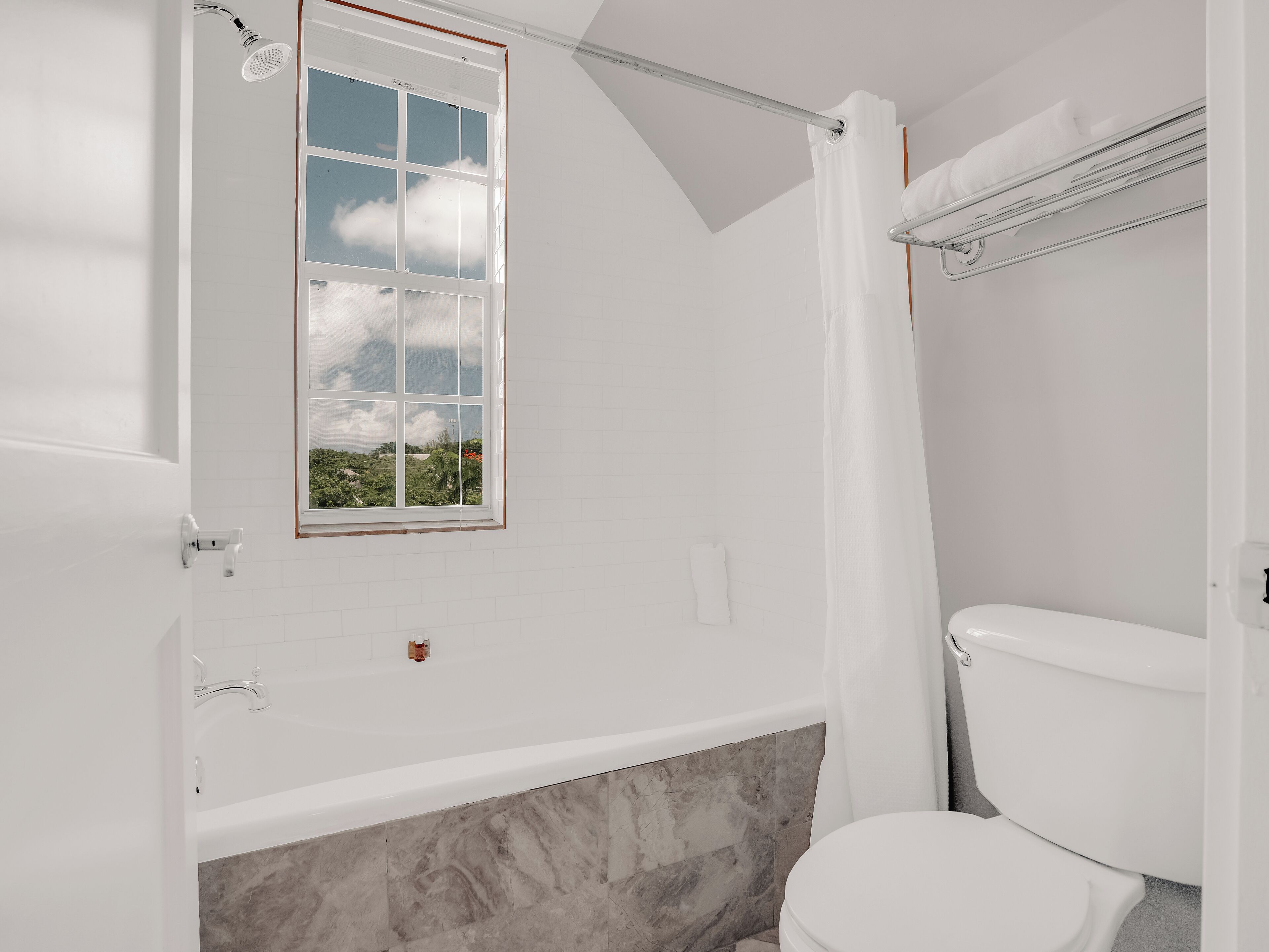 Et badeværelse med badekar, toilet, et vindue med udsigt over grøn vegetation og en bruseforhæng.