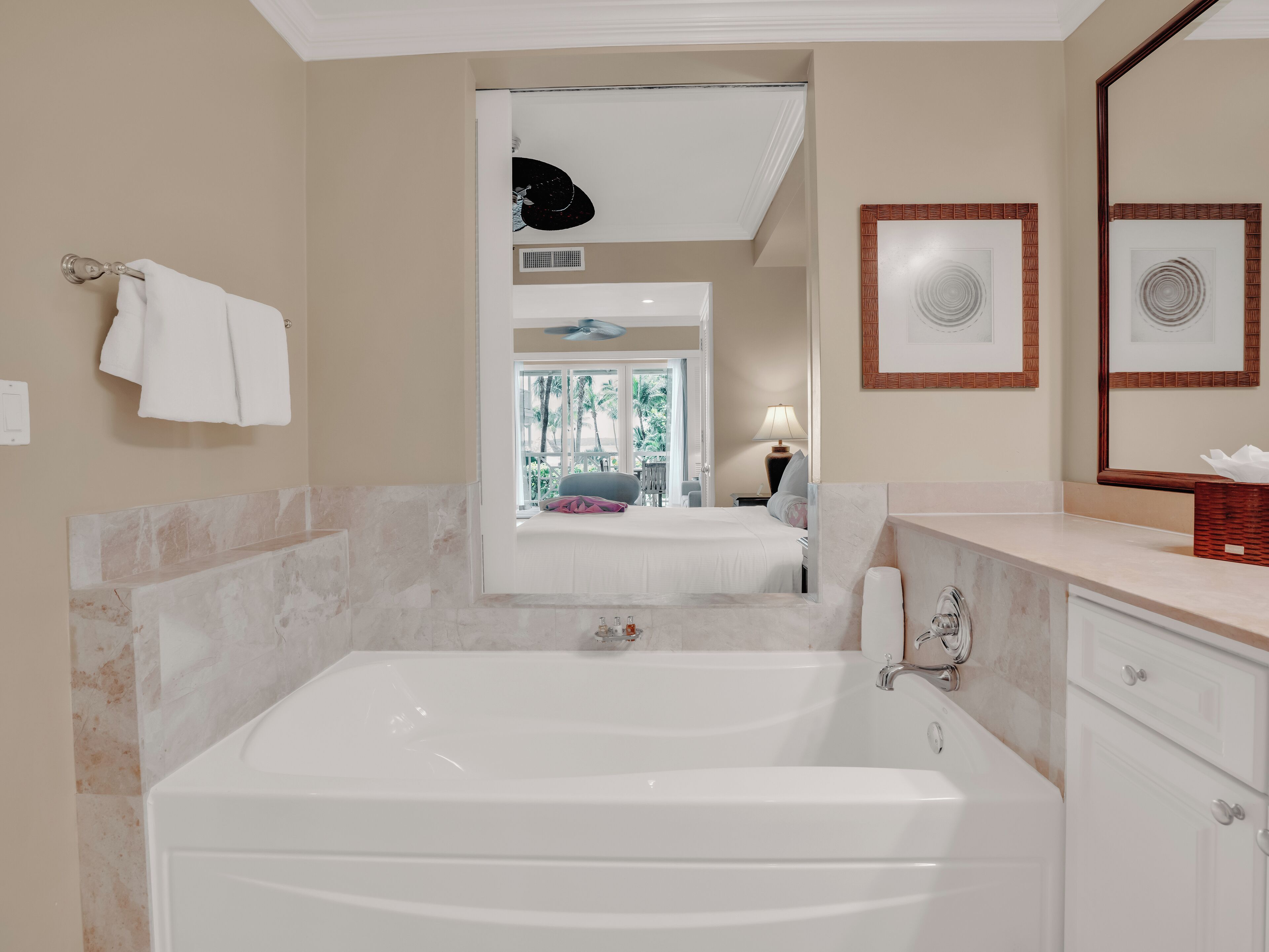 A modern bathroom with a large bathtub, a vanity with a mirror, and a view of a bedroom with a bed and a window.