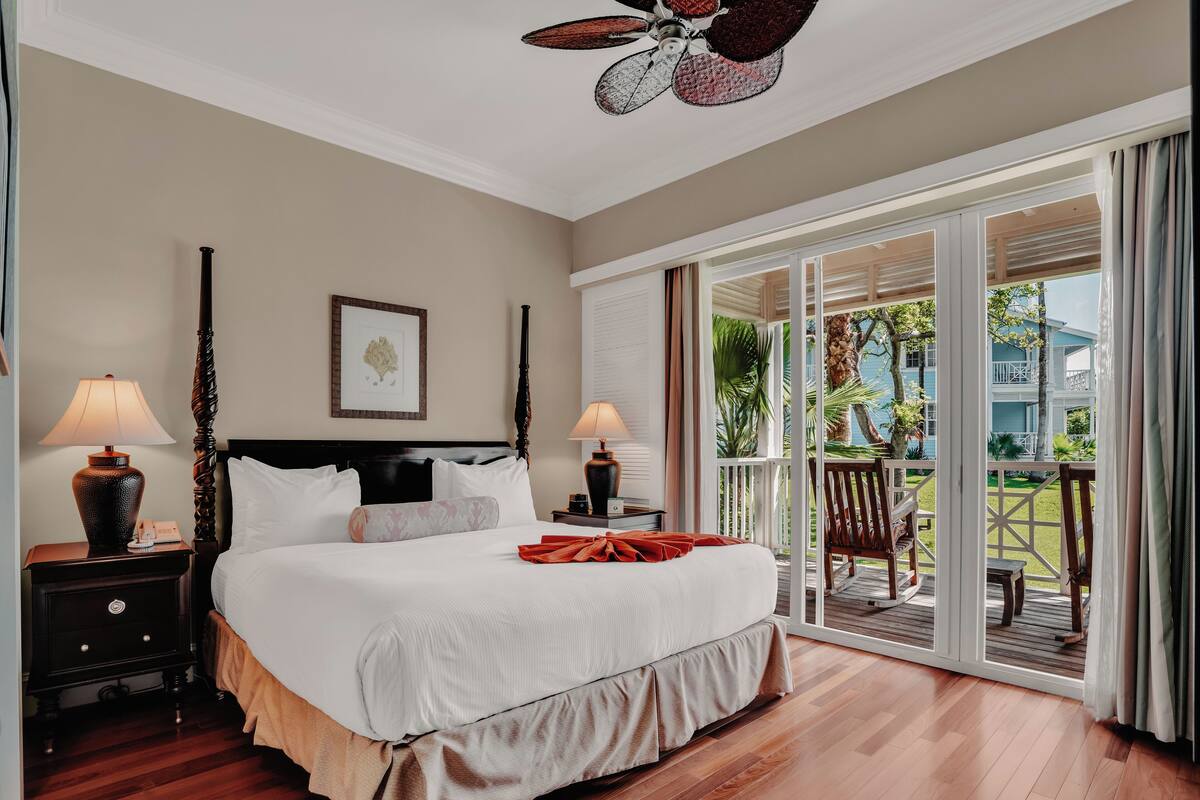 A hotel room with a large bed, two bedside tables with lamps, a ceiling fan, and a view of a pool and building through a sliding glass door.