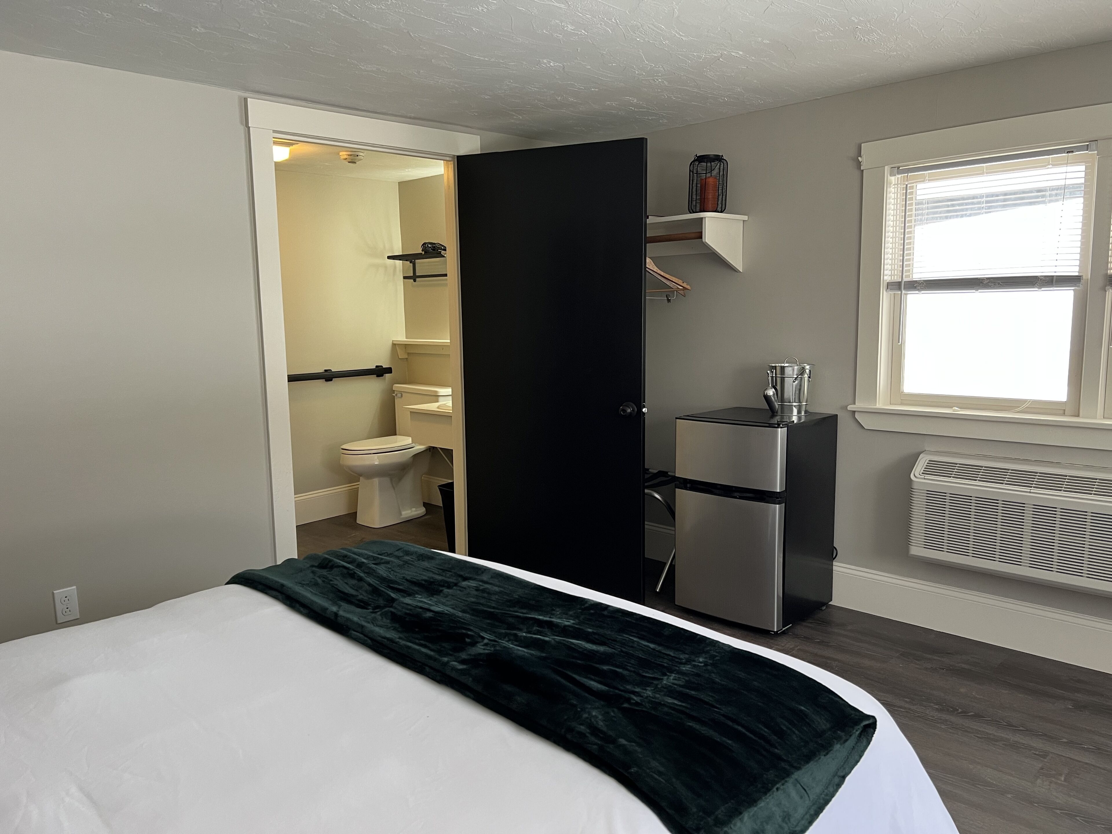 Un dormitorio con una cama, un armario negro, una pequeña kitchenette y un baño visible a través de una puerta abierta.