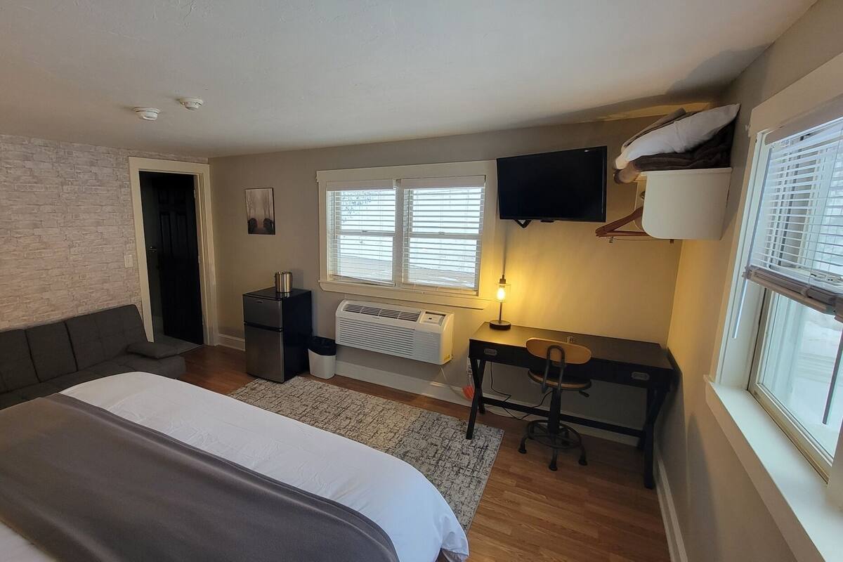 A bedroom with a bed, a desk, a chair, a heater, a TV, and a window with blinds.
