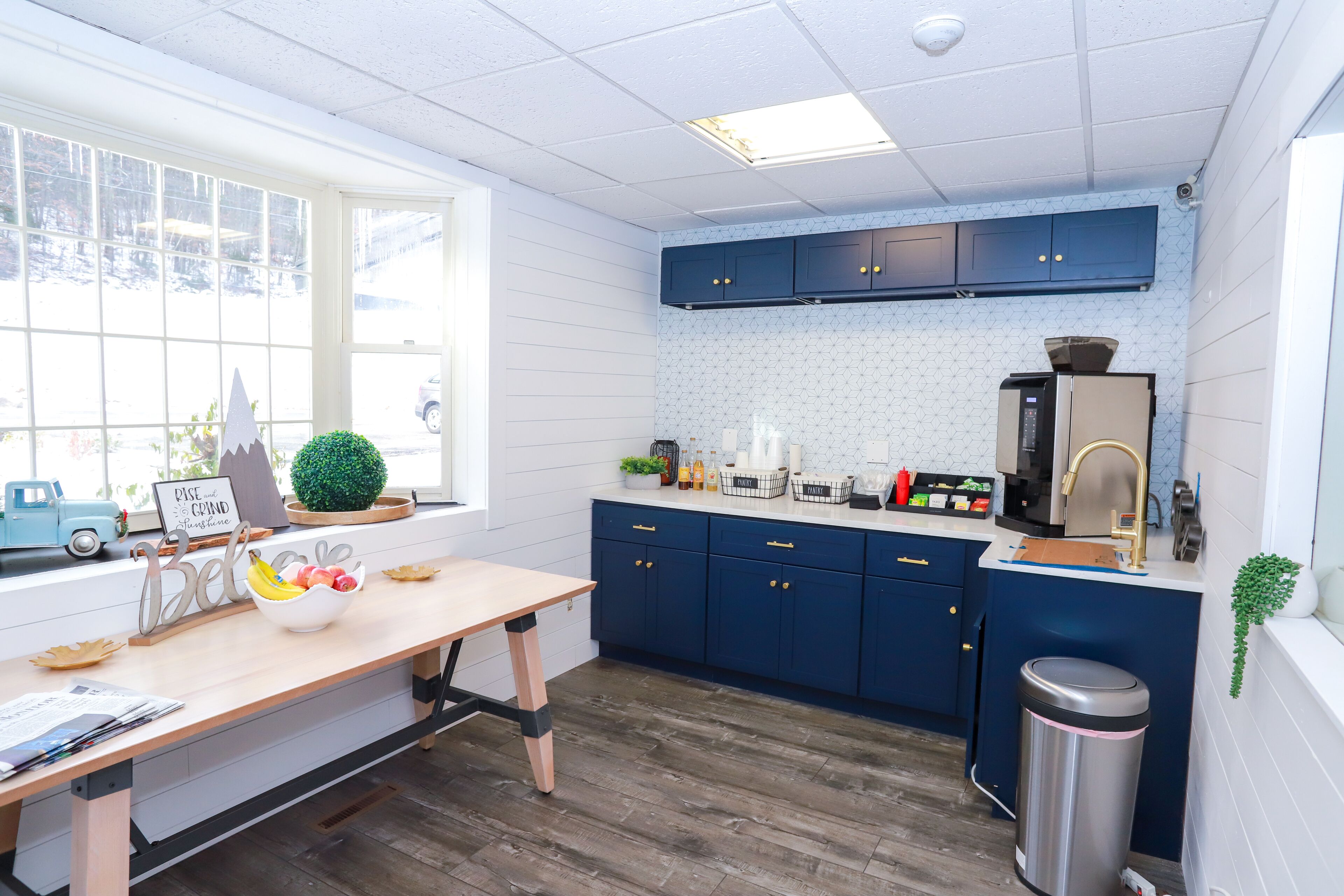 A kitchen with wooden flooring, a window with a view of a snowy landscape, and a countertop with various appliances and a trash can.