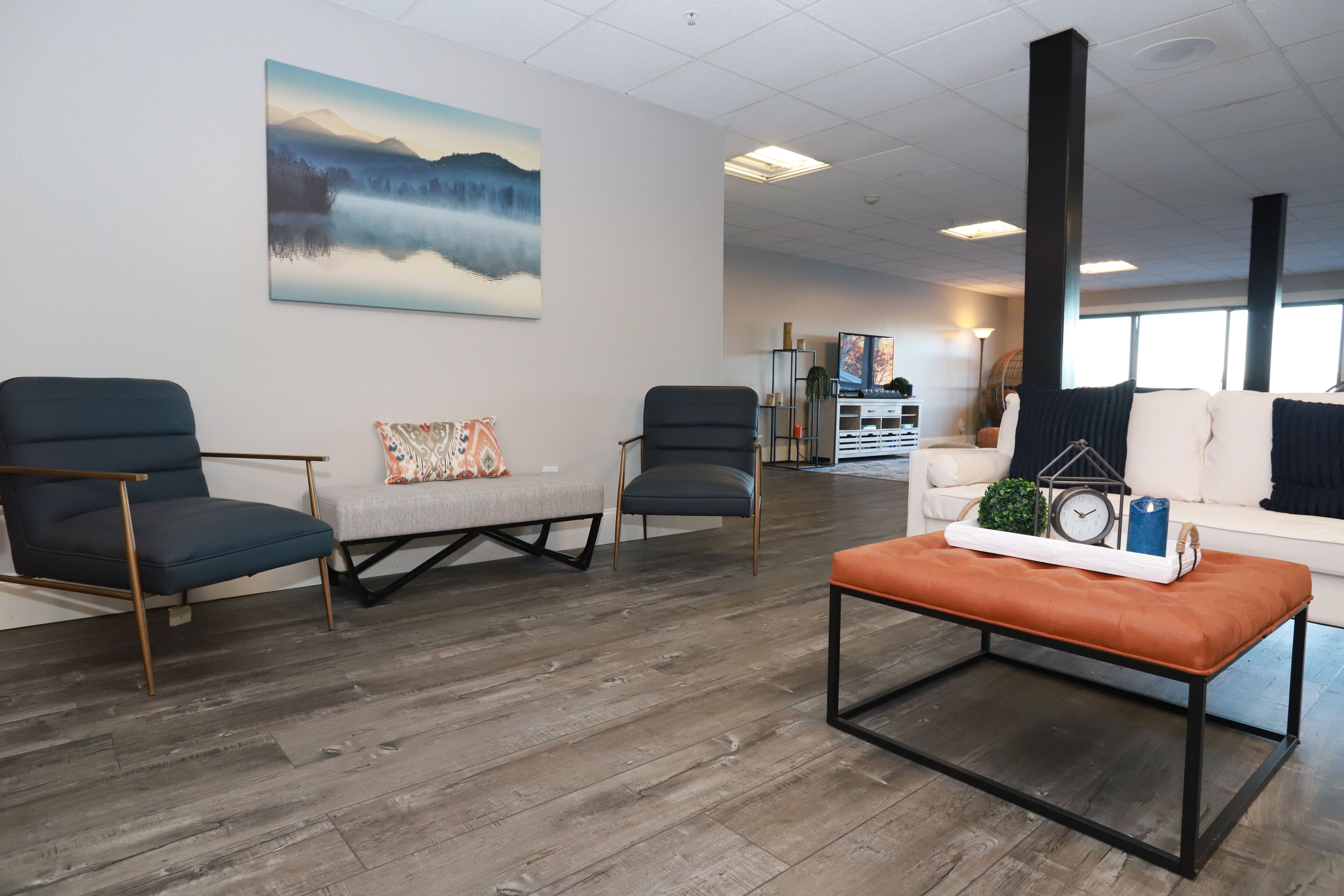 A modern living room with a wooden floor, two armchairs, a sofa, and a coffee table. A painting of a mountain landscape is on the wall.