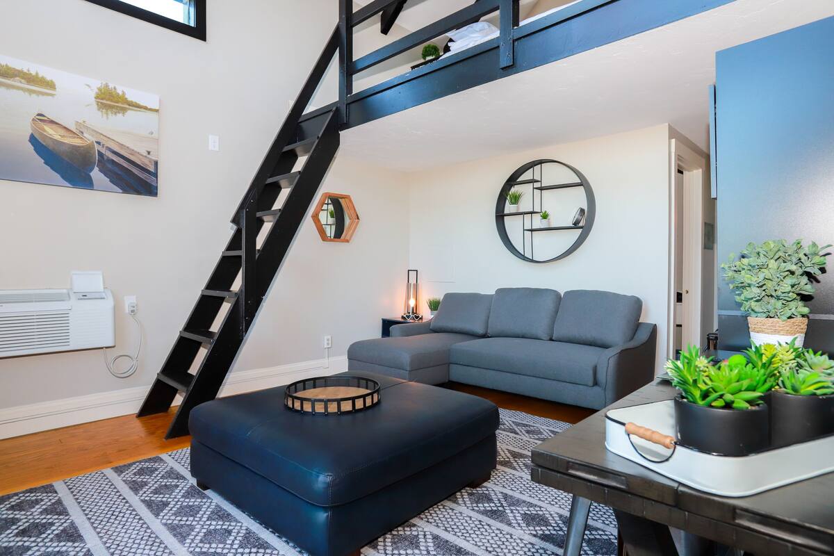A modern living room with a gray sectional sofa, a black ottoman, a round mirror, and a staircase.