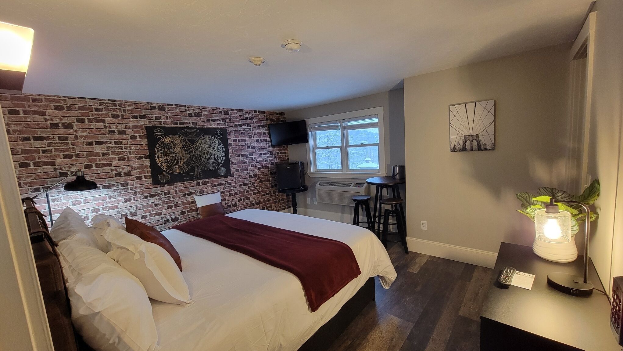 A hotel room with a brick wall, a bed with white linens, a desk with a lamp, and a window with a view.