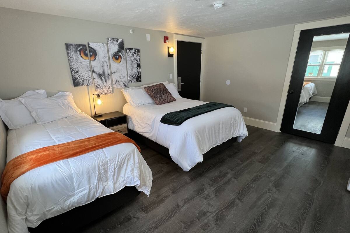 A hotel room with two beds, a nightstand, a wall-mounted lamp, and a door leading to a bathroom.