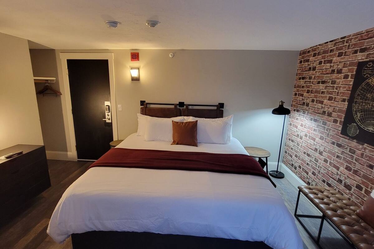 A hotel room with a bed, a desk, a lamp, a door, and a brick wall with a globe artwork.