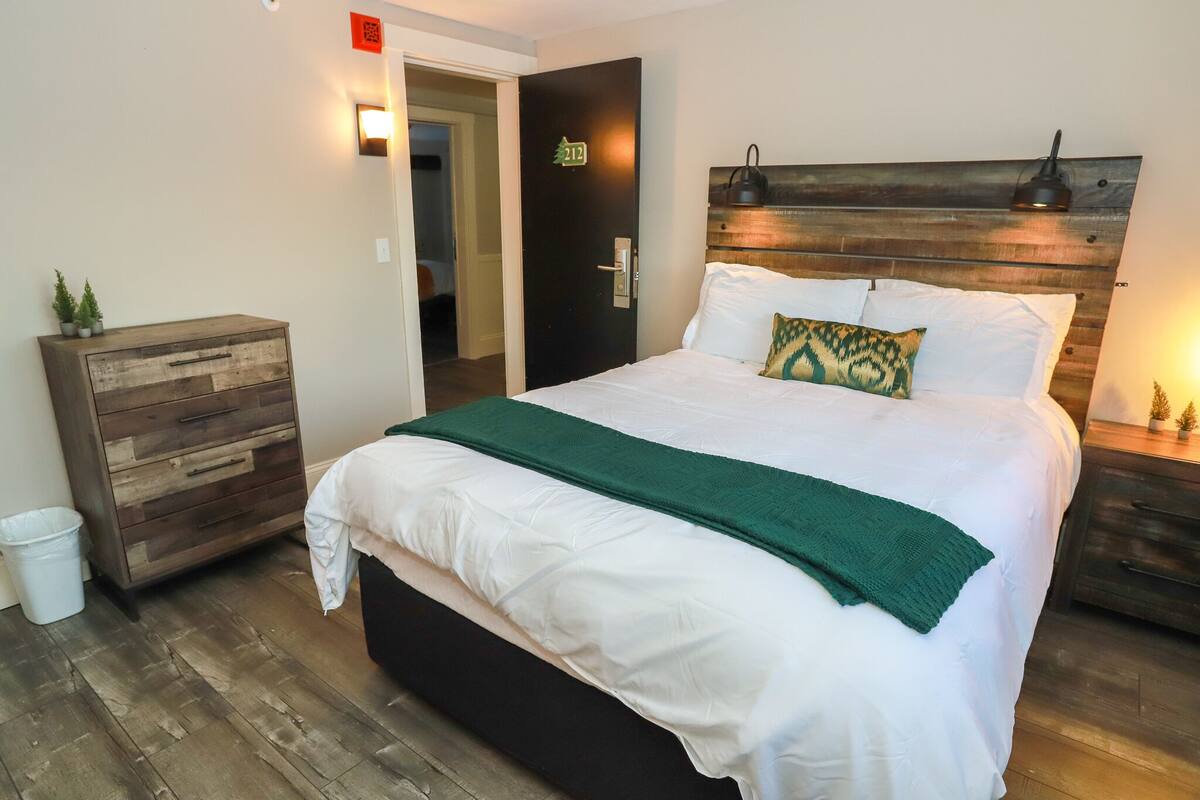 A neatly made bed with a green throw, a wooden nightstand with a lamp, and a wooden dresser with a trash bin.