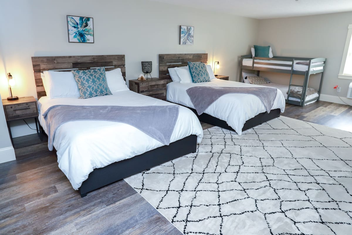 A modern bedroom with two beds, a bunk bed, wooden headboards, bedside tables with lamps, and abstract wall art.