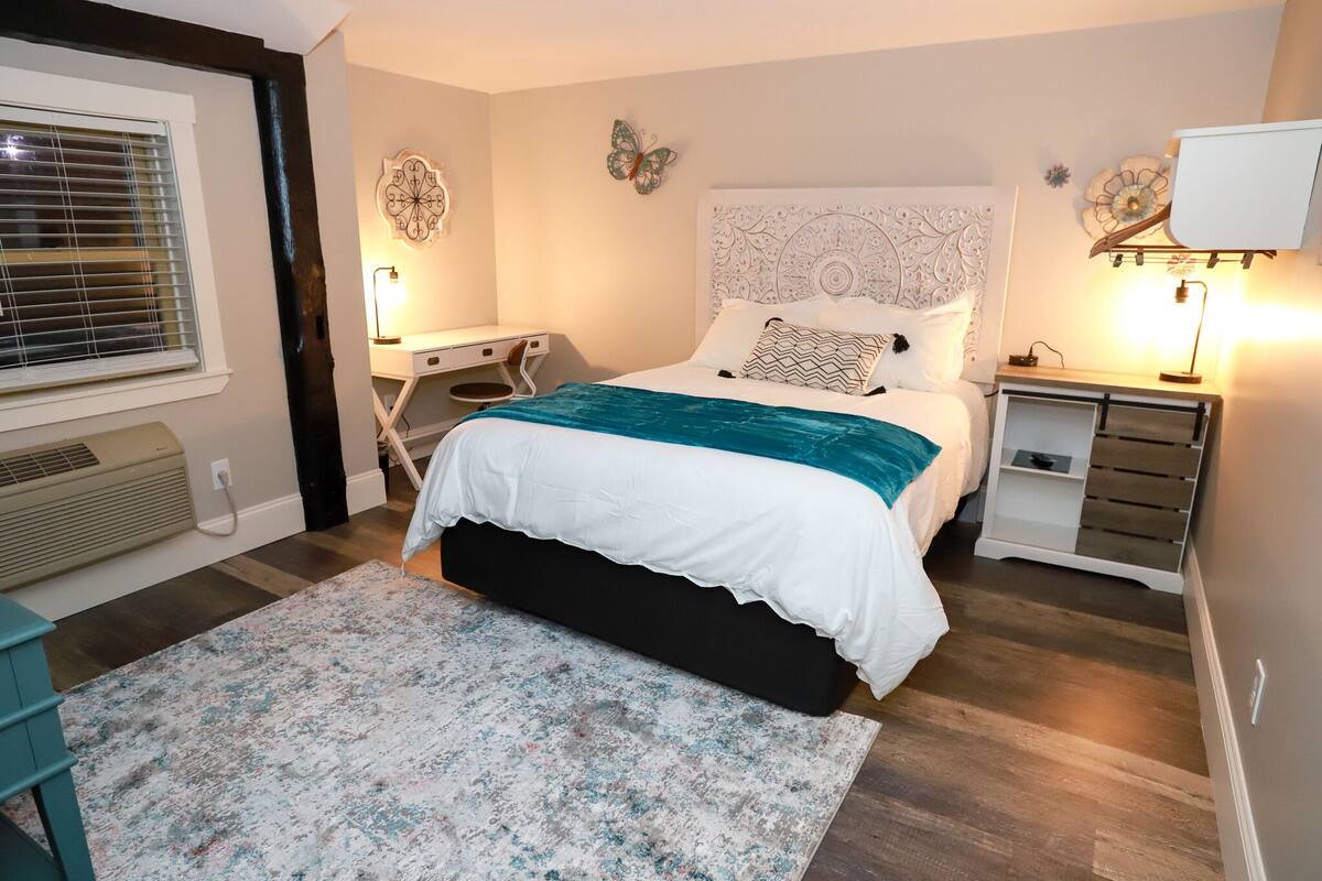 A bedroom with a bed, bedside tables, a desk, and a window with blinds.