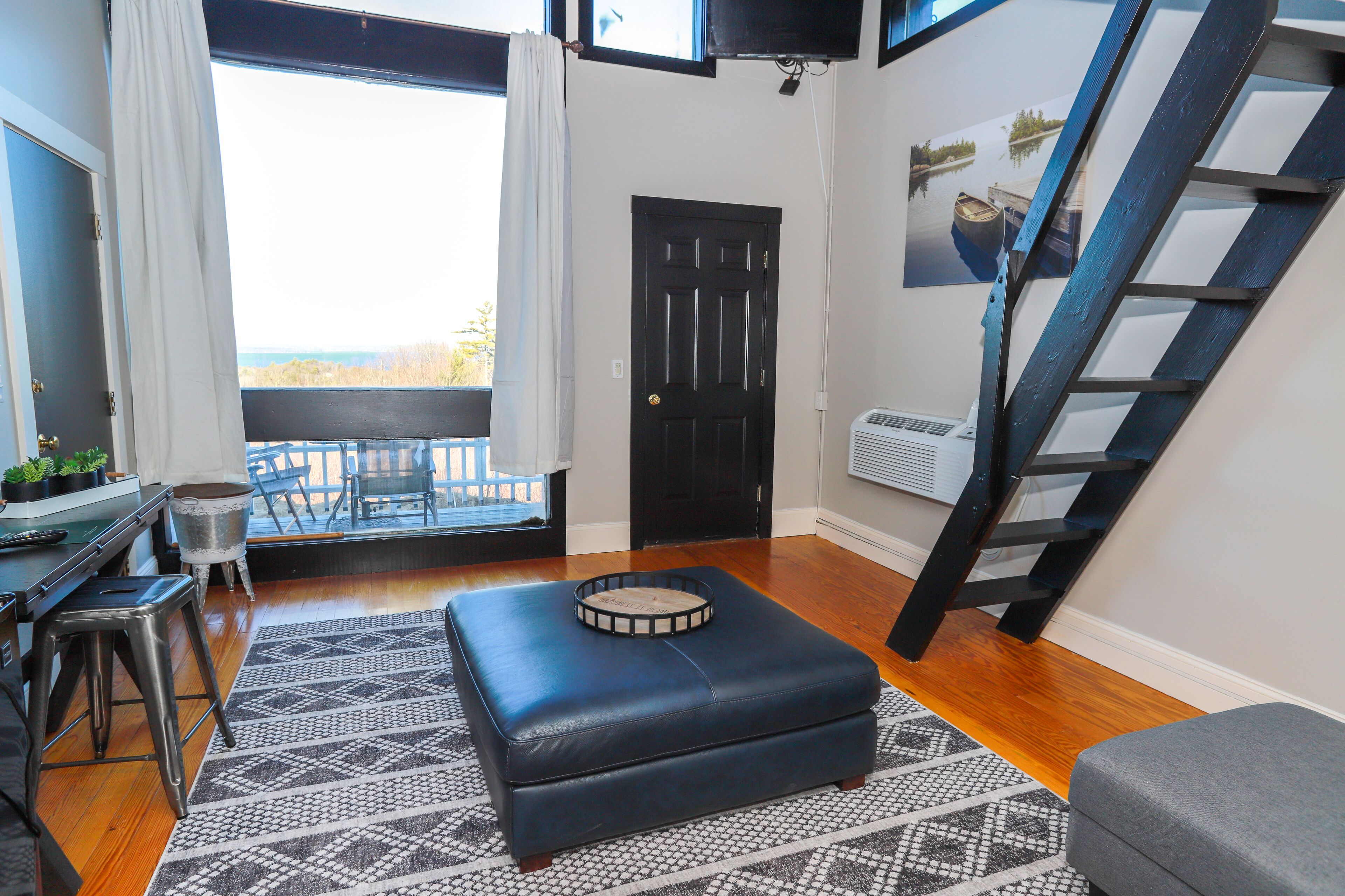 A room with a black door, a window with white curtains, a black and white patterned rug, a black ottoman, a grey bench, and a black ladder leaning against the wall.