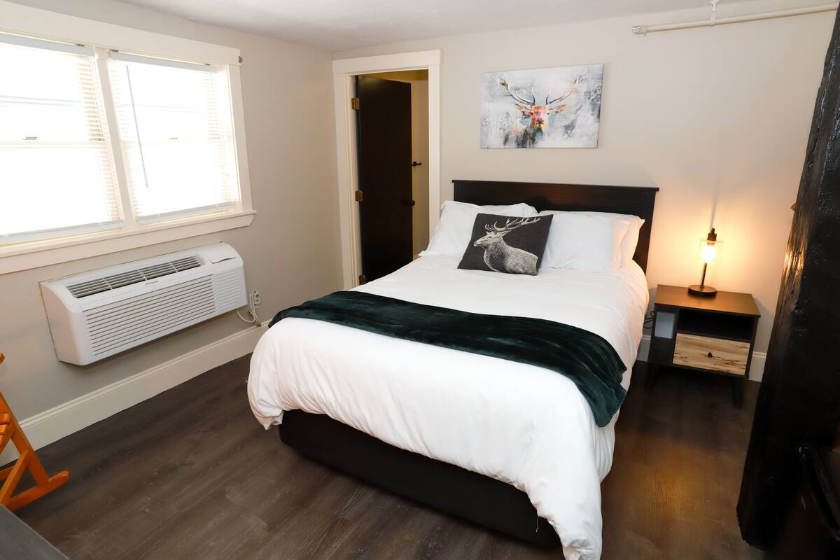 A bedroom with a bed, a nightstand, a lamp, a window with blinds, and an air conditioning unit.