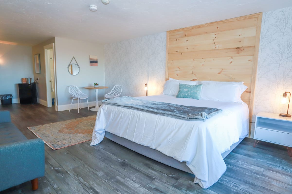 A bedroom with a wooden headboard, a bed with white bedding, a small desk with a chair, and a nightstand with a lamp.