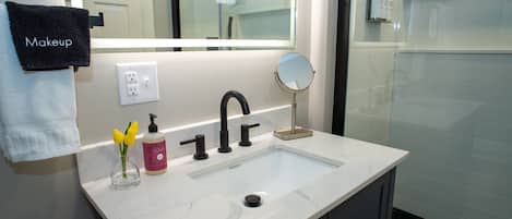 A bathroom with a white sink, black faucet, and a mirror. There is a towel rack with a black towel labeled "Makeup" and a small vase with yellow flowers.