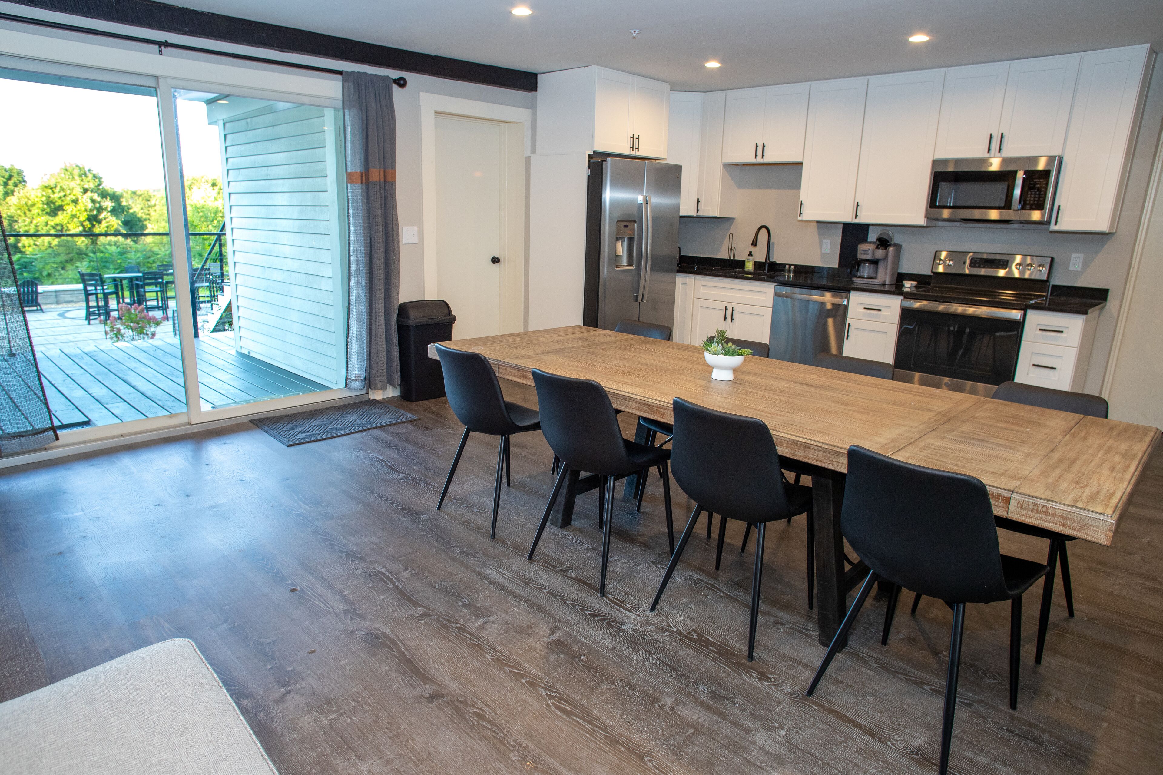A modern kitchen with a large wooden table, black chairs, white cabinets, stainless steel appliances, and a sliding glass door leading to an outdoor deck.