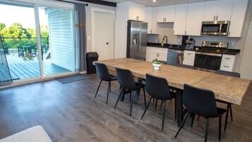 A modern kitchen with a large wooden table, black chairs, white cabinets, stainless steel appliances, and a sliding glass door leading to an outdoor deck.