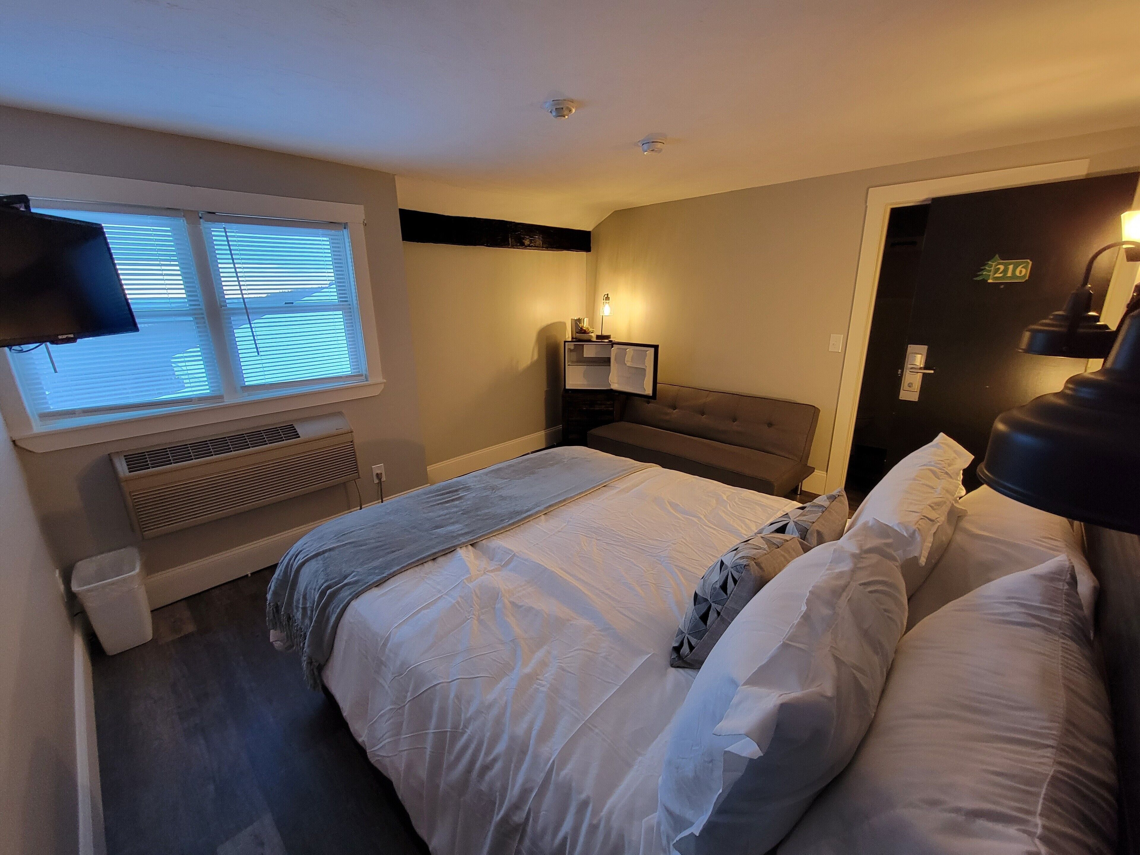 Habitación de hotel con una cama grande, un televisor montado en la pared, una ventana con persianas, una mesita de noche con lámpara y una puerta con el número.
