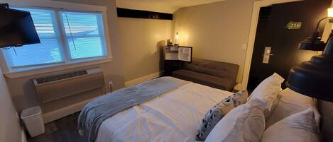 Habitación de hotel con una cama grande, un televisor montado en la pared, una ventana con persianas, una mesita de noche con lámpara y una puerta con el número.