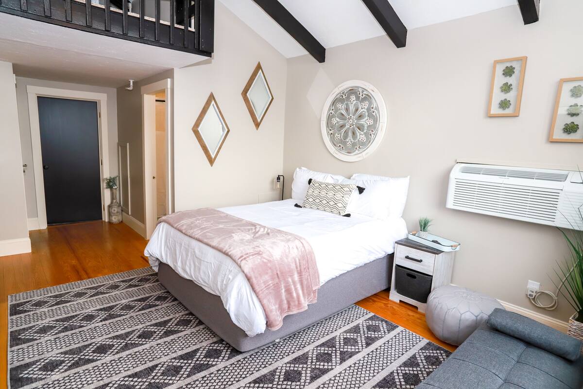 A bedroom with a bed, bedside tables, a nightstand, a wall-mounted air conditioner, and a window with blinds.