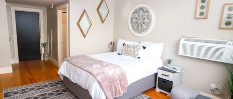 A bedroom with a bed, bedside tables, a nightstand, a wall-mounted air conditioner, and a window with blinds.