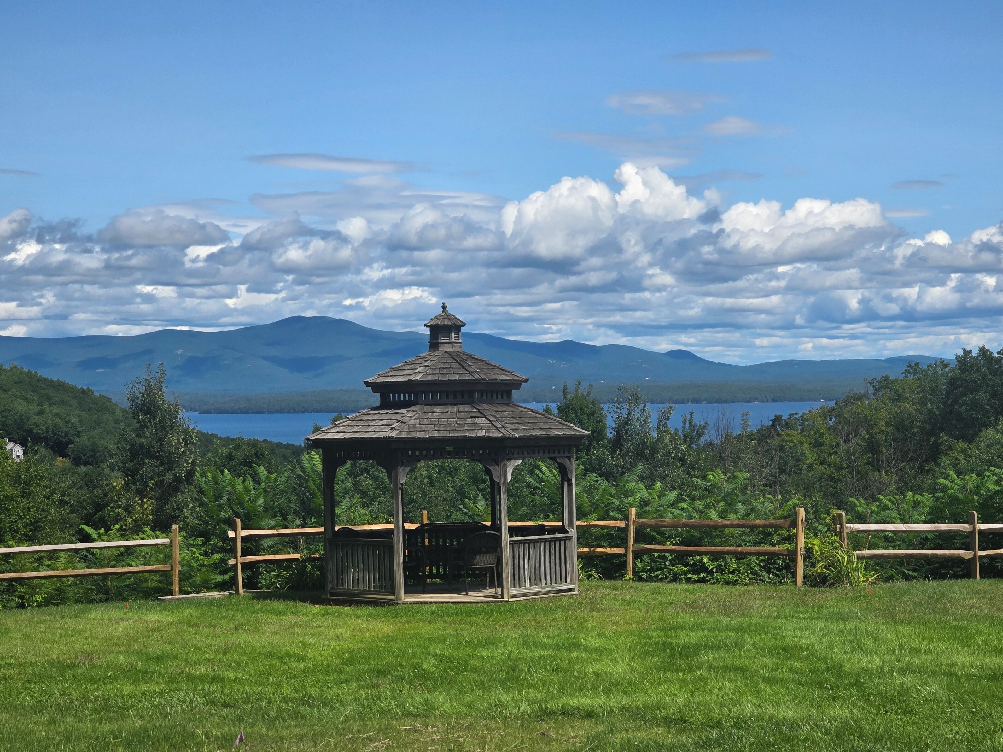 A gazebo in a grassy field with a lake and mountains in the background.