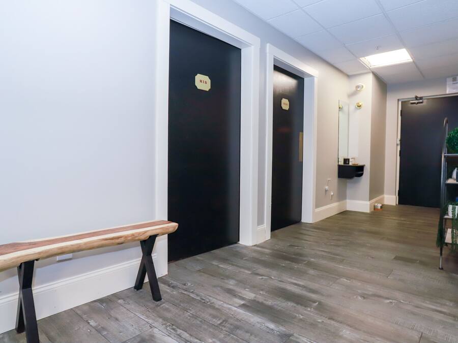 Un couloir avec un banc, des portes noires, un miroir et une étagère sur laquelle sont posés divers objets.
