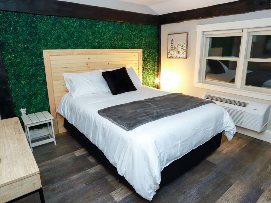 Een slaapkamer met een houten hoofdbord, een bed met wit beddengoed en een zwart kussen, een klein nachtkastje, een groene muur, een raam met zonwering, en een verwarming.