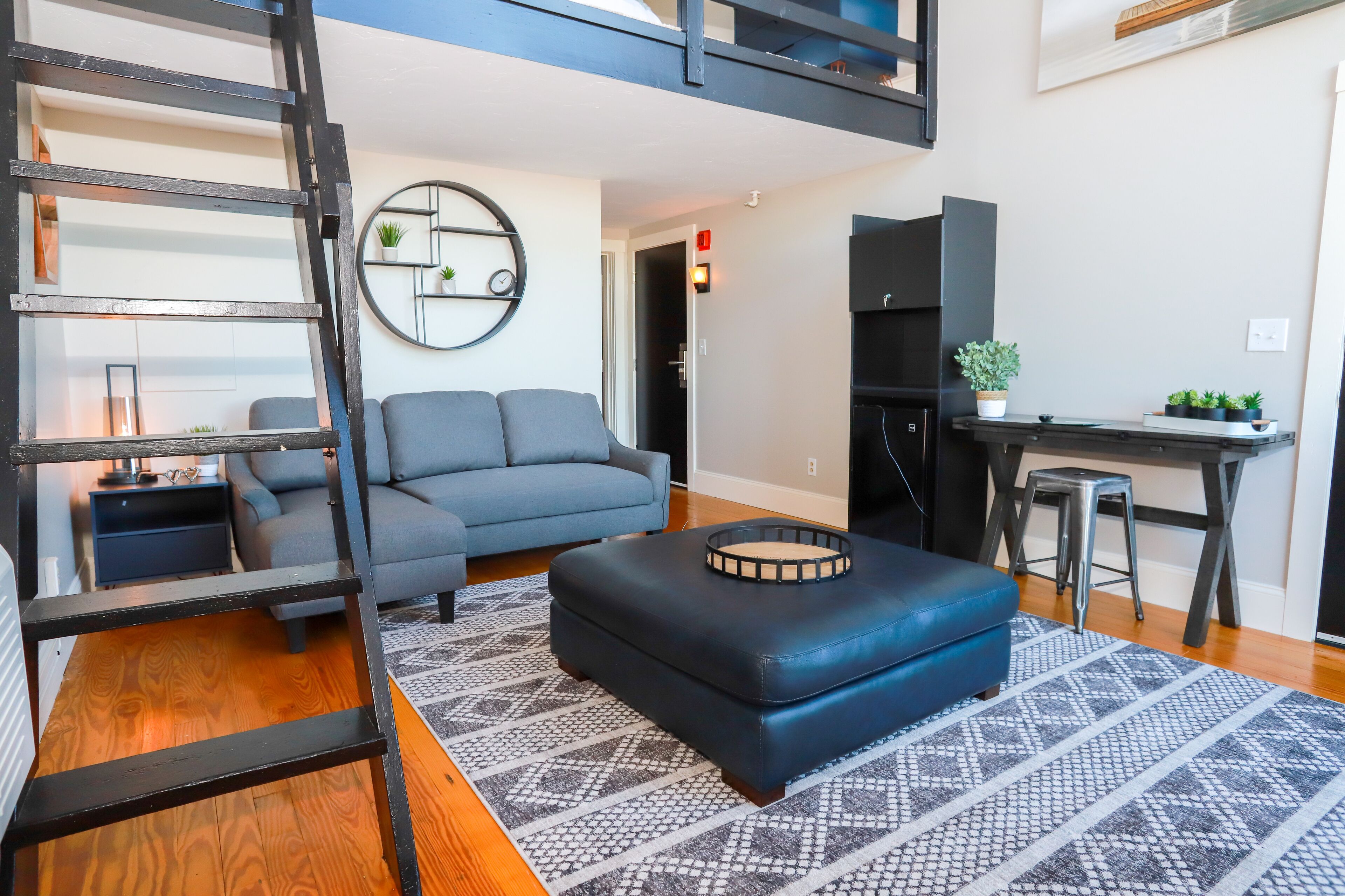 A modern living room with a grey sofa, a black ottoman, a patterned rug, and a black ladder.